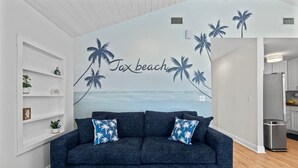 A living room with a dark blue sofa, two blue pillows with palm tree patterns, a white shelf unit with decorative items, and a wall mural depicting a beach scene with palm trees and the text "Tax beach". - Endless Summer Retreat (Jacksonville Beach)