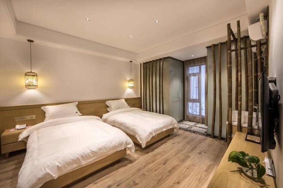 A hotel room with two beds, wooden flooring, a desk with a plant, and a large window with curtains.