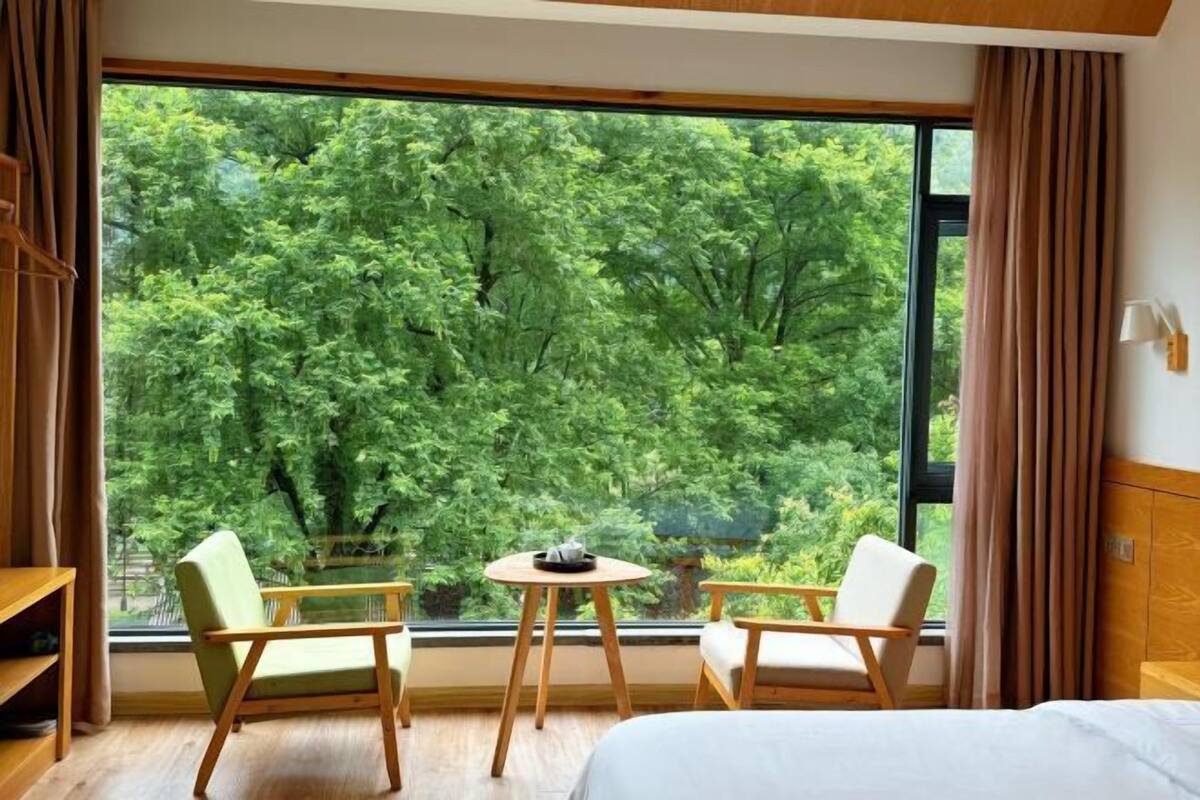 A room with a bed, two chairs, a small table, and a large window with a view of trees.