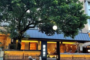 Un restaurant japonès tradicional amb façana de fusta, un arbre gran al davant i un teulat de palla.
