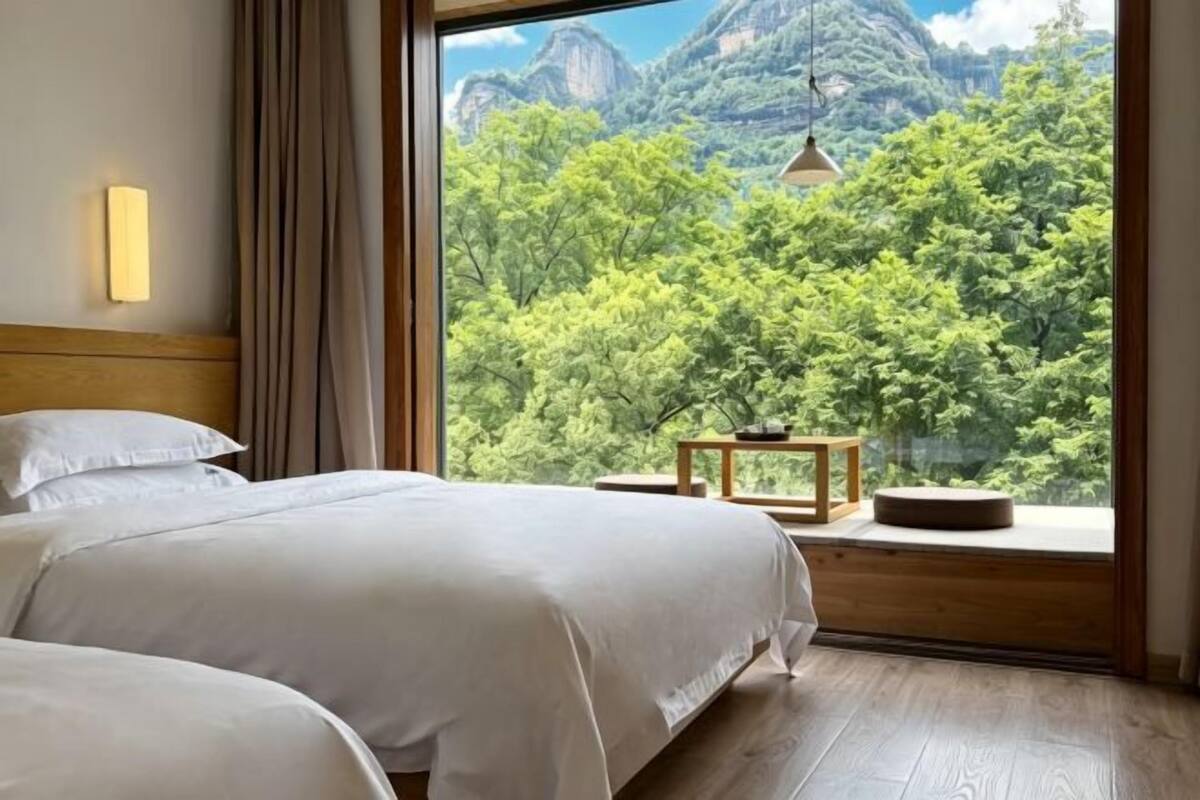 A hotel room with two beds, a wooden headboard, a large window with a mountain view, and a small table with a lamp.