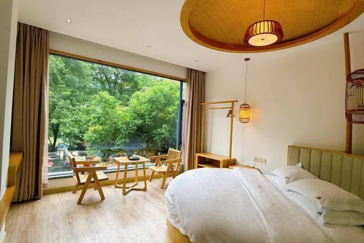 A bedroom with a large window, a bed, a table with chairs, and a hanging light fixture.