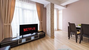 Un saló modern amb un televisor de pantalla plana que mostra 'NETFLIX', una taula de dinar de fusta amb cadirees i una finestra gran amb cortines primes.