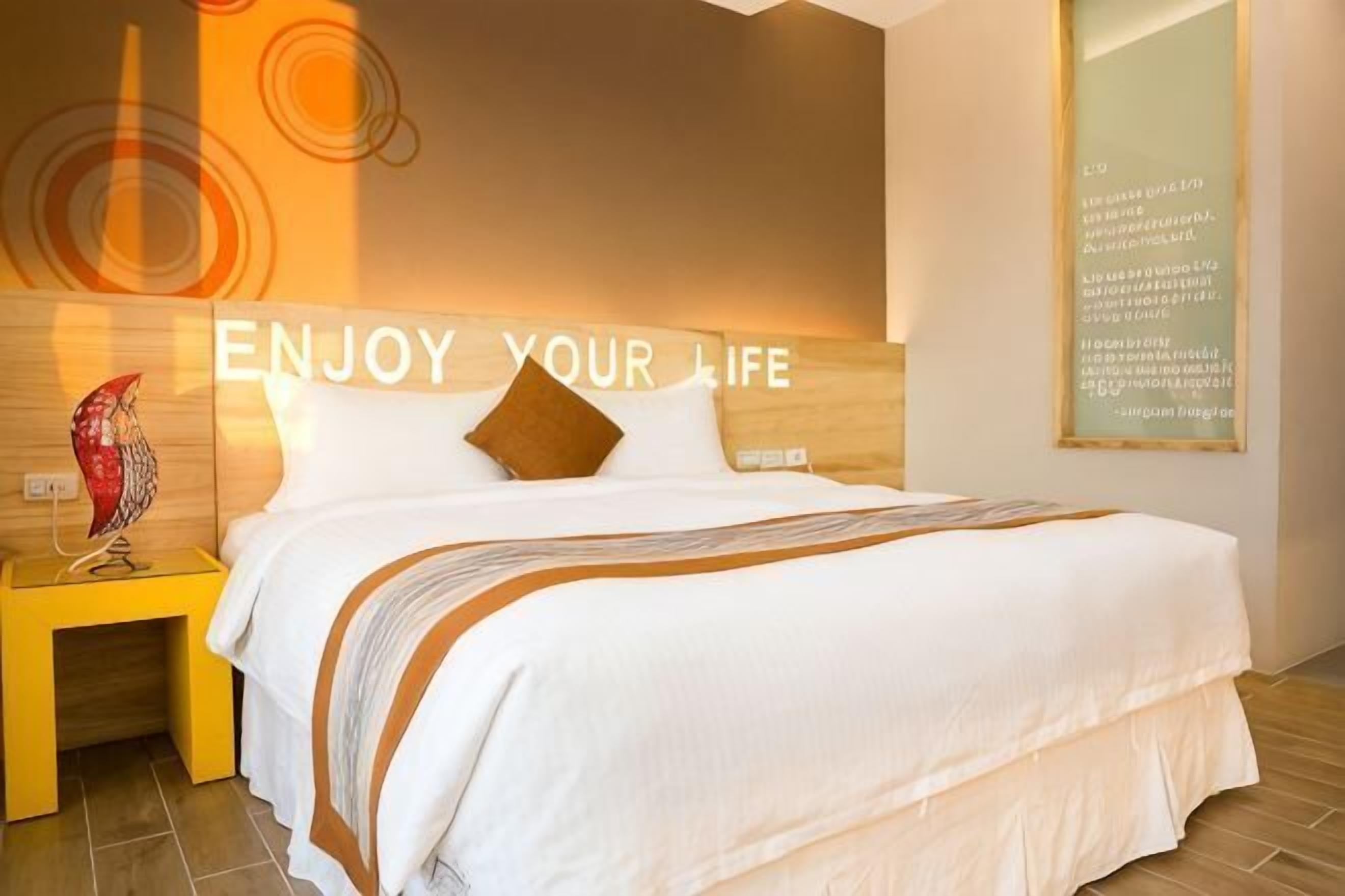 A neatly made bed with a headboard, a bedside table with a lamp, and a wall with a motivational quote.
