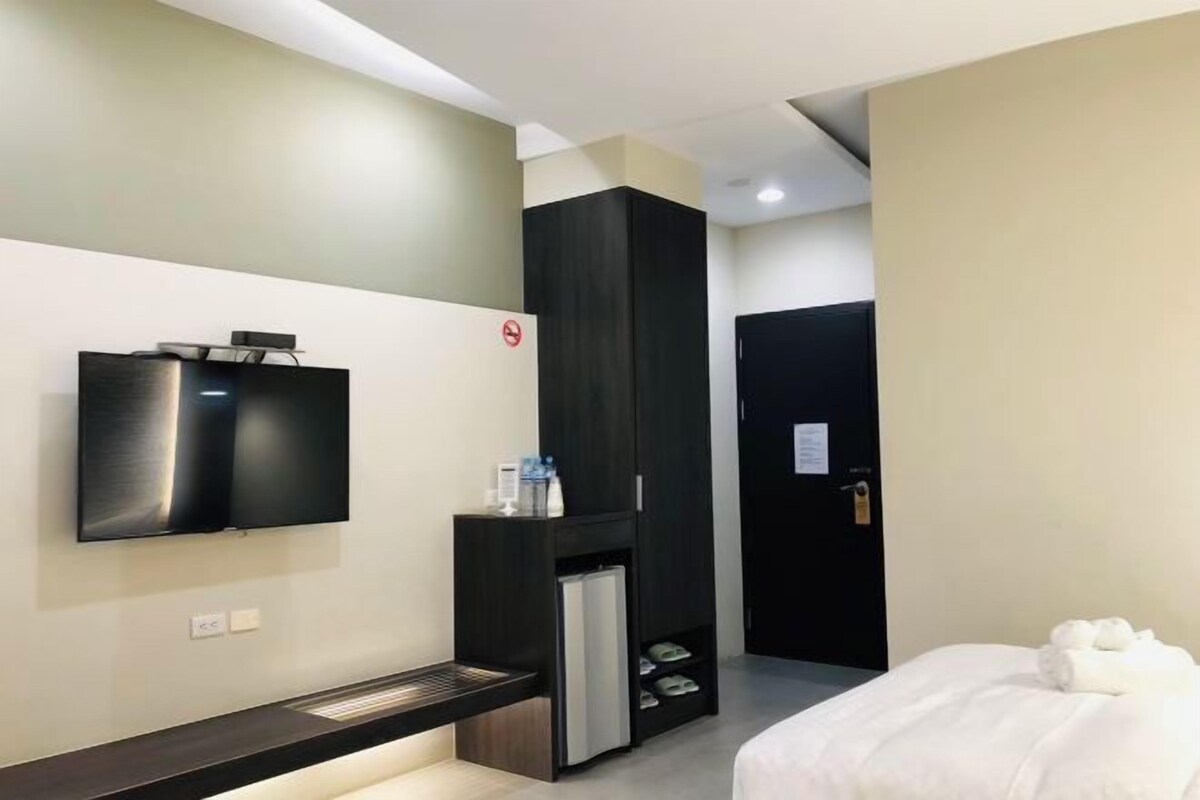 A modern hotel room with a flat-screen TV mounted on the wall, a built-in mini-fridge, a black wardrobe, and a bed with white bedding.