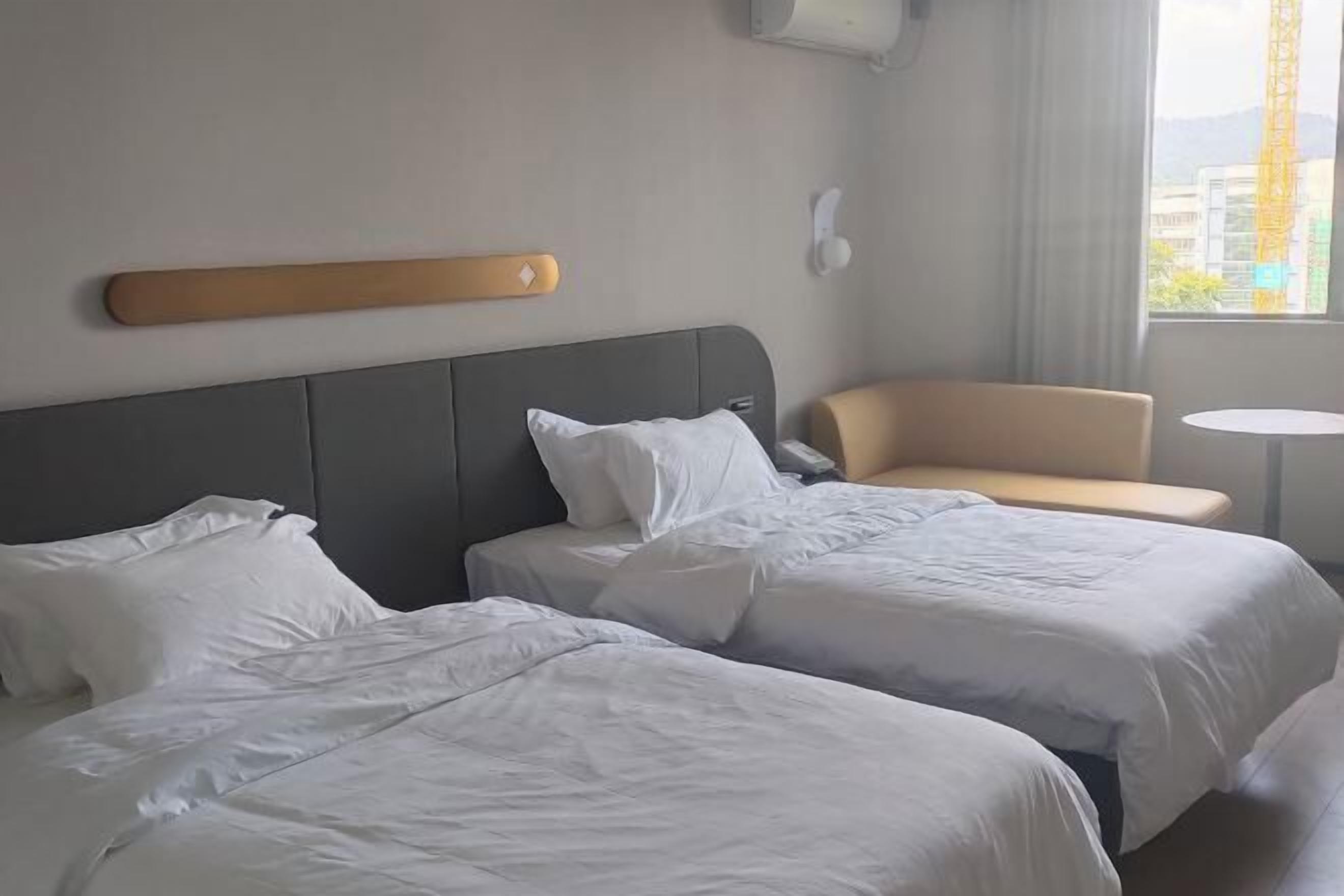 A hotel room with two beds, a small sofa, a round table, and a window with a view of buildings and a crane.