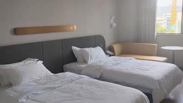 A hotel room with two beds, a small sofa, a round table, and a window with a view of buildings and a crane.
