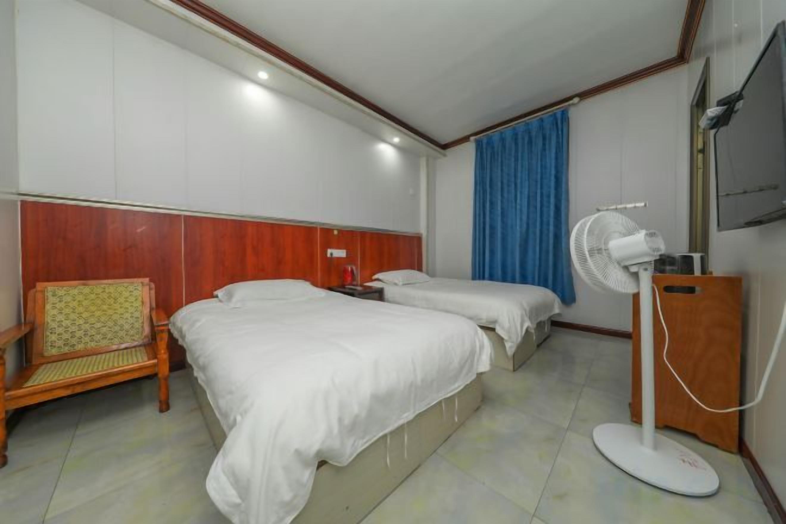 A hotel room with two beds, a wooden headboard, a desk, a chair, a standing fan, and a TV.