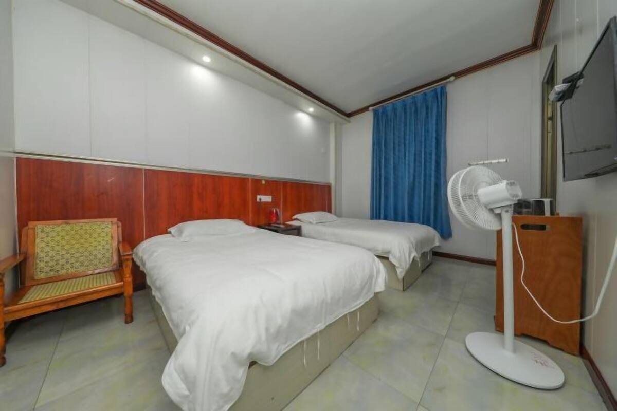 Ein Hotelzimmer mit zwei Betten, einem Holzstuhl, einem Standventilator und einem an der Wand befestigten Fernseher.