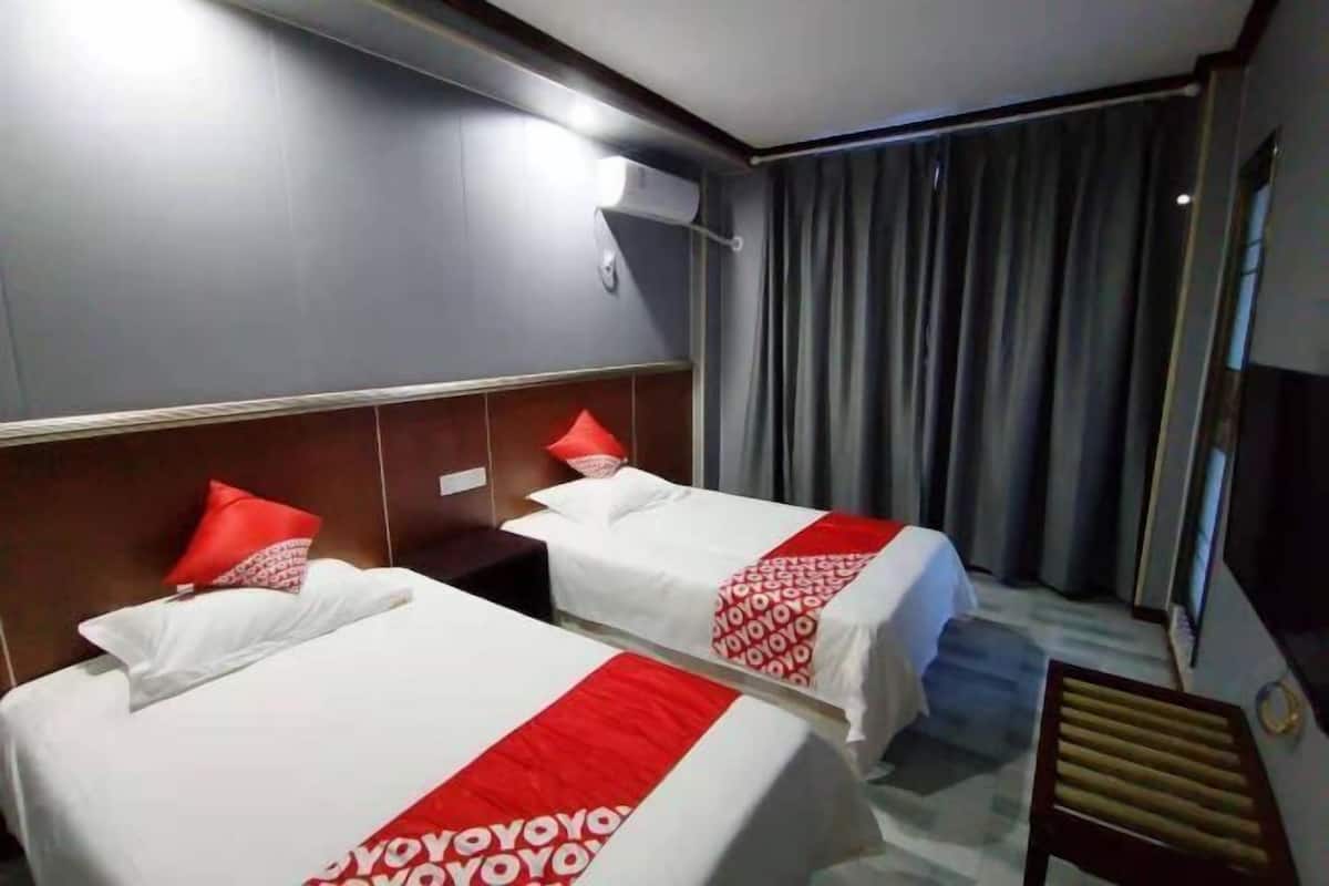 Ein Hotelzimmer mit zwei Betten, jeweils mit einer rot-weißen Tagesdecke, einem Kopfteil, einem Nachttisch und einem Fenster mit Vorhängen.