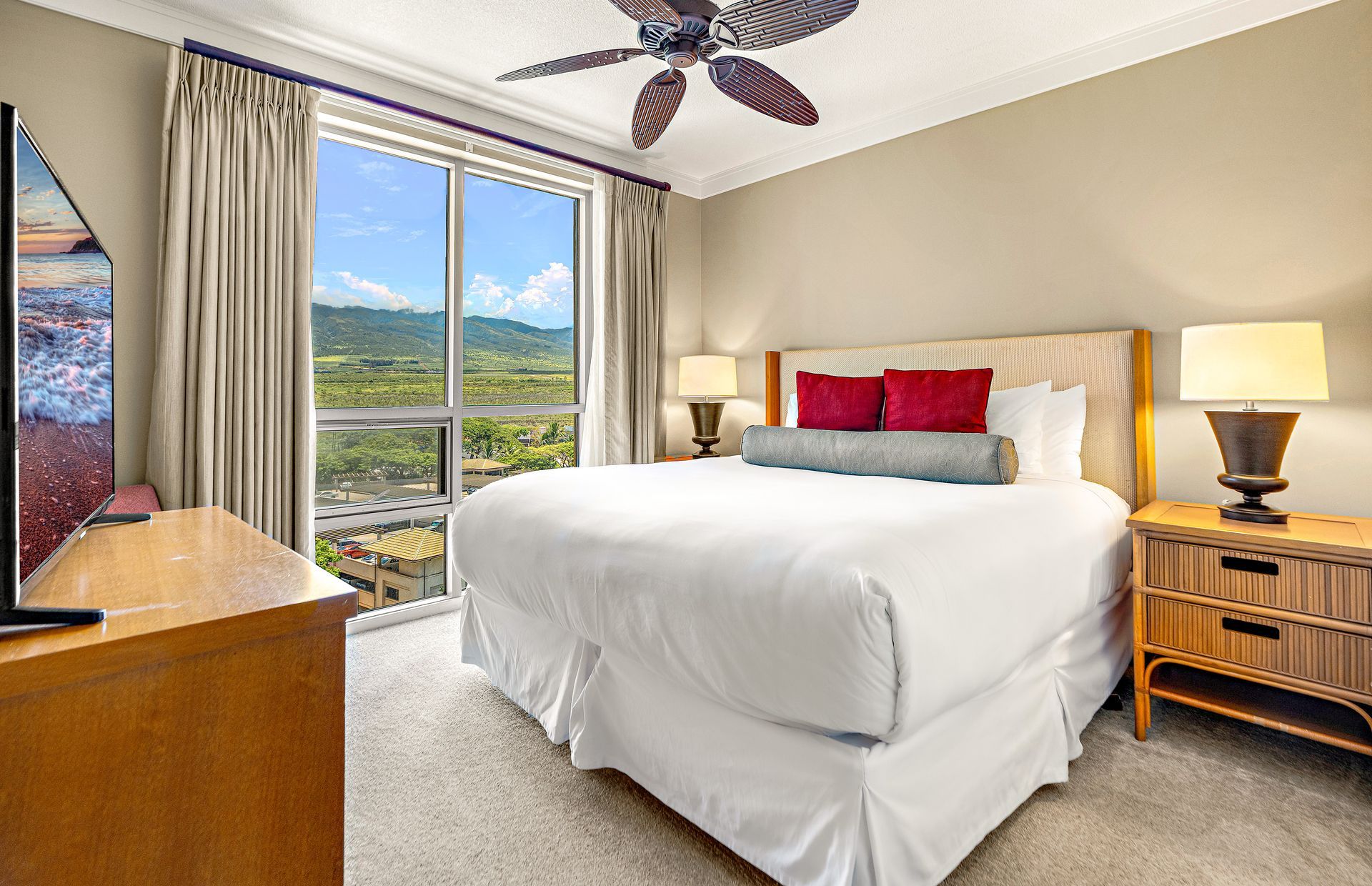 Une chambre d’hôtel avec un grand lit, deux lampes de chevet, un ventilateur de plafond, une télévision et une vue sur un paysage par la fenêtre.