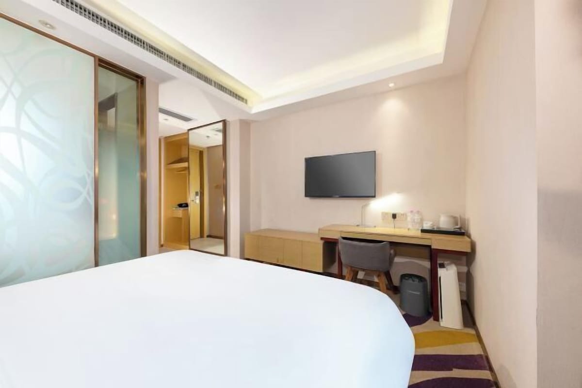 A hotel room with a bed, a desk with a chair, a television, and a trash bin.