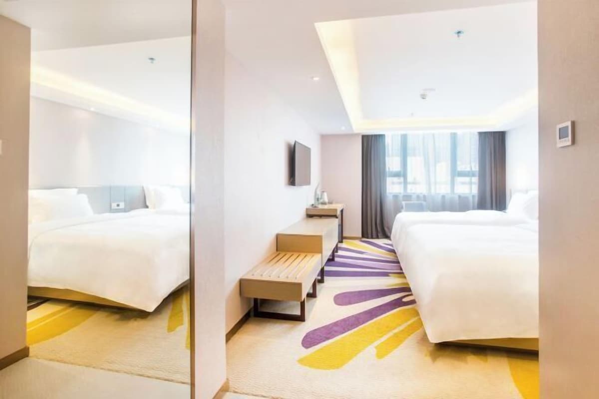 A hotel room with two beds, a bench, a small table, and a large window.
