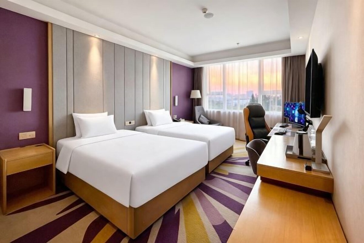 Ein Hotelzimmer mit zwei Betten, einem Schreibtisch, einem Stuhl, einem Fernseher und einem Fenster mit Blick auf die Stadt.