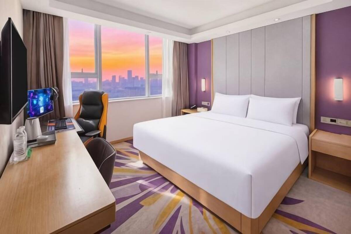 Ein Hotelzimmer mit einem großen Bett, einem Schreibtisch mit Computer, einem Sessel und Blick auf die Skyline der Stadt.