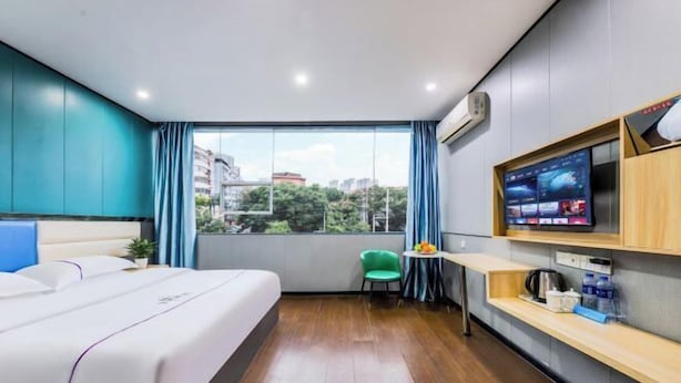 A hotel room with a large bed, a small table with a chair, a TV mounted on the wall, and a window with a view of buildings and trees. - Yijia Hotel (Ji'an No.2 Middle School Pedestrian Street) (Ji'an)