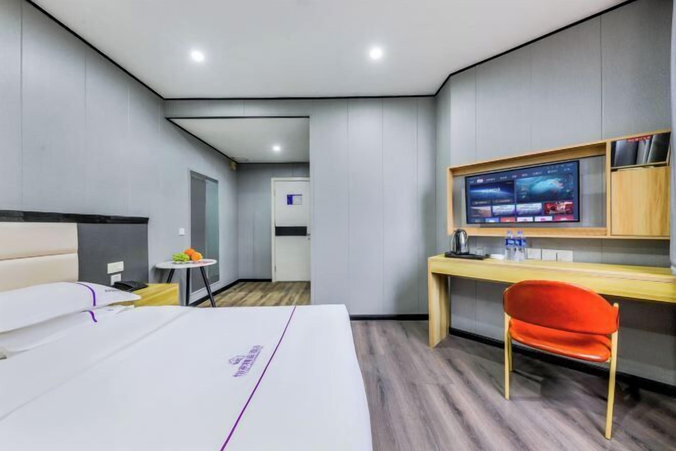 A hotel room with a bed, a desk with a computer monitor, a chair, and a small table with a fruit bowl.