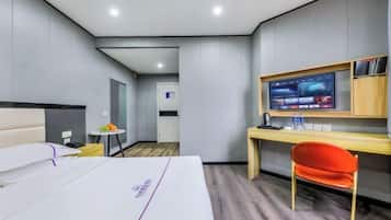 A hotel room with a bed, a desk with a computer monitor, a chair, and a small table with a fruit bowl.