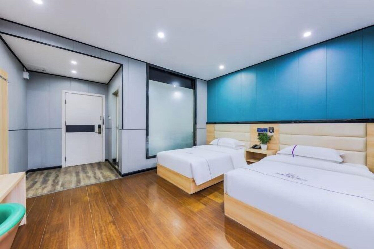 A modern hotel room with two beds, a wooden floor, a blue accent wall, and a small bedside table with a plant.