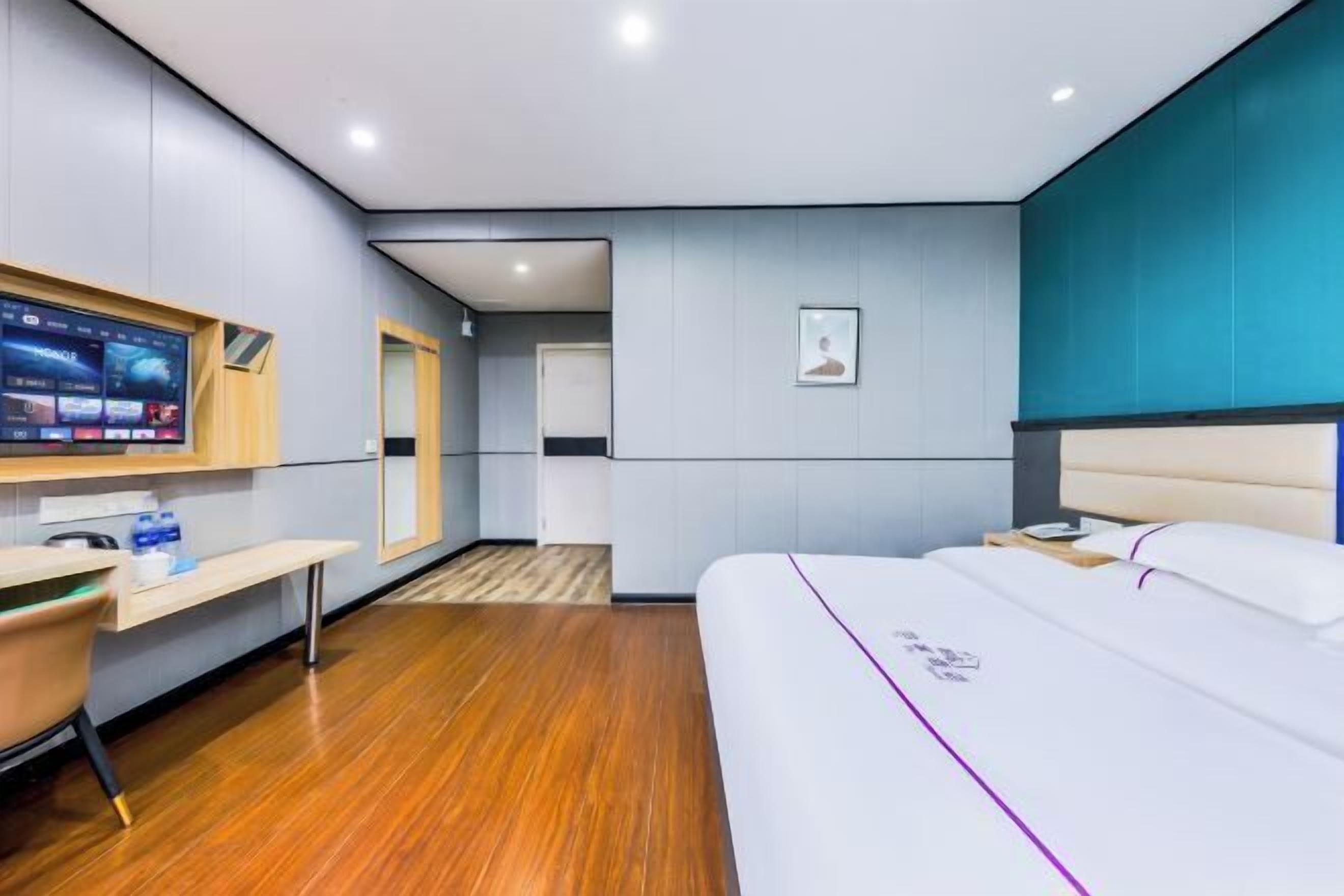 A modern hotel room with a wooden floor, a bed with a white headboard, a desk with a chair, a TV mounted on the wall, and a framed picture on the wall.