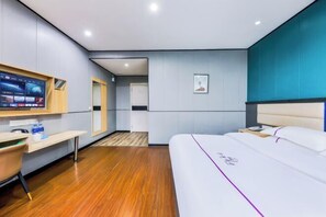 A modern hotel room with a wooden floor, a bed with a white headboard, a desk with a chair, a TV mounted on the wall, and a framed picture on the wall.