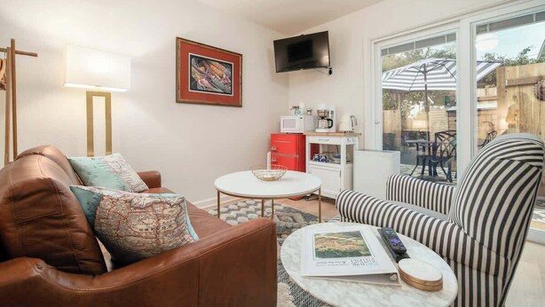 Un salon avec un canapé en cuir marron, un fauteuil à rayures, une table basse blanche, un téléviseur à écran plat fixé au mur et une fenêtre donnant sur l’extérieur, où se trouve un parapluie.