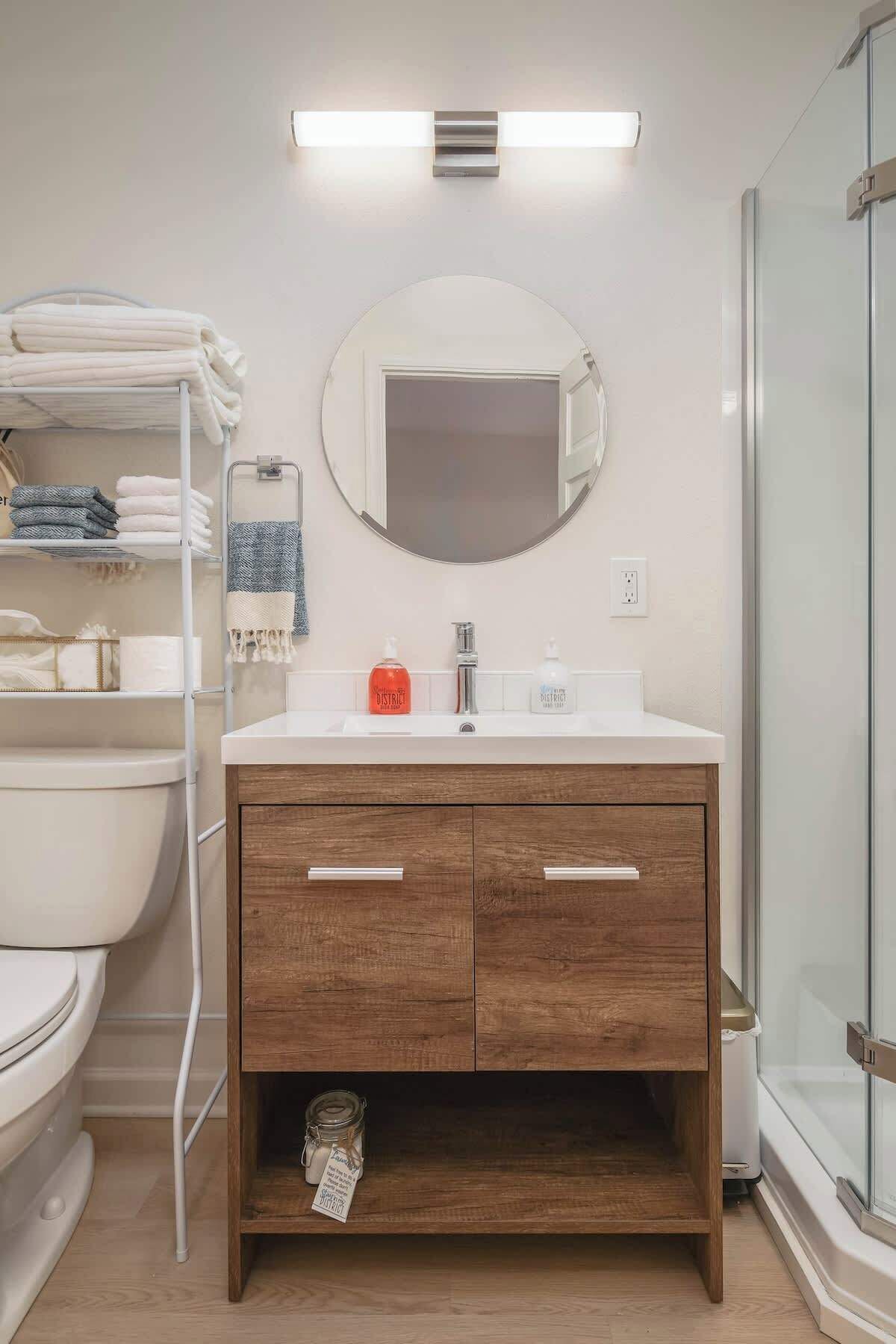 A bathroom with a white toilet, a wooden vanity with two drawers, a round mirror, and a glass shower enclosure.