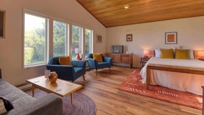 A spacious room with a large bed, a seating area with blue chairs, a small table with a vase, and large windows offering a view of trees. - #StayinMyDistrict Cape Arago Studio Suite (Coos Bay)