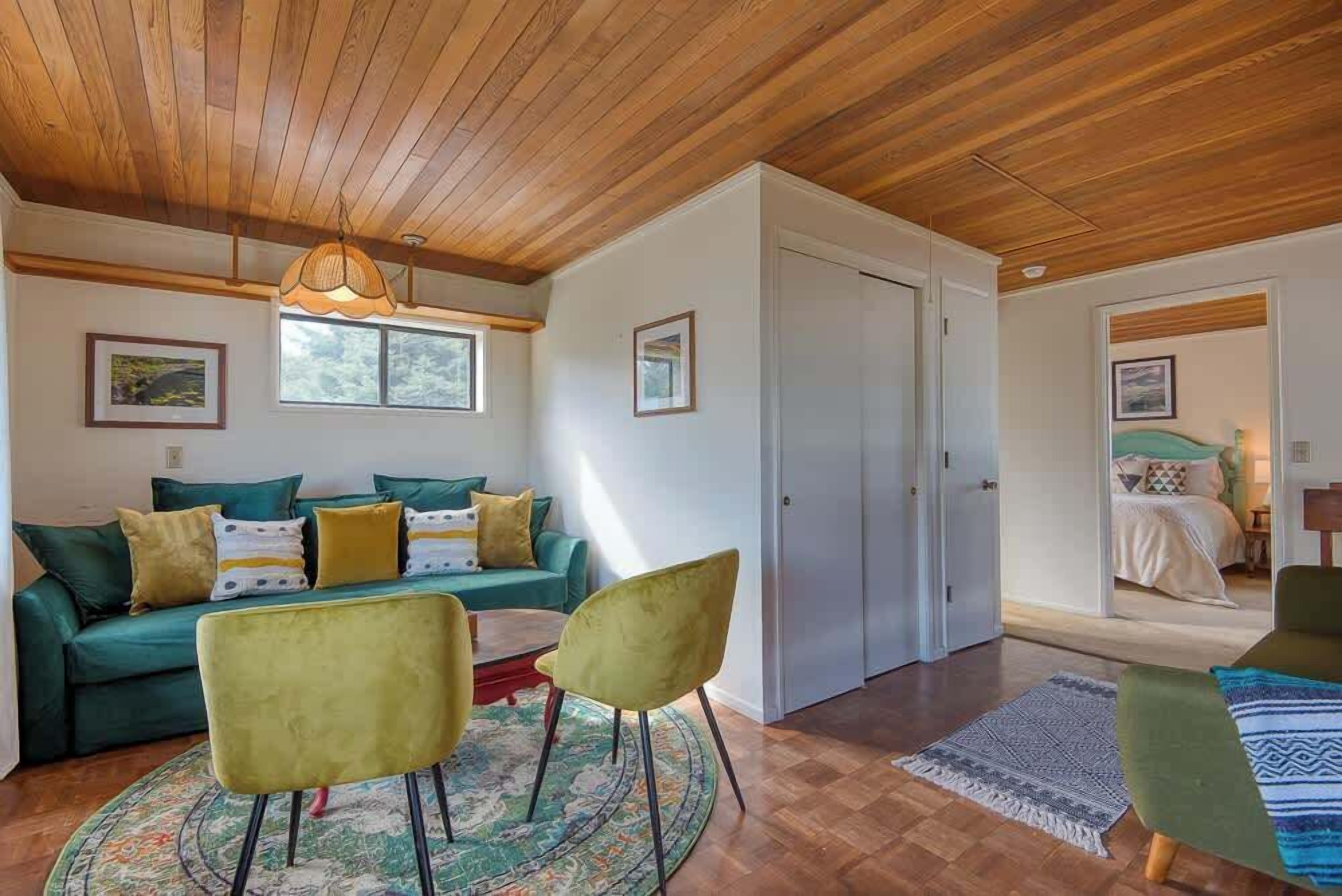 Un salon chaleureux avec un plafond en bois, un canapé vert agrémenté de coussins jaunes, deux fauteuils verts et un tapis à motifs.