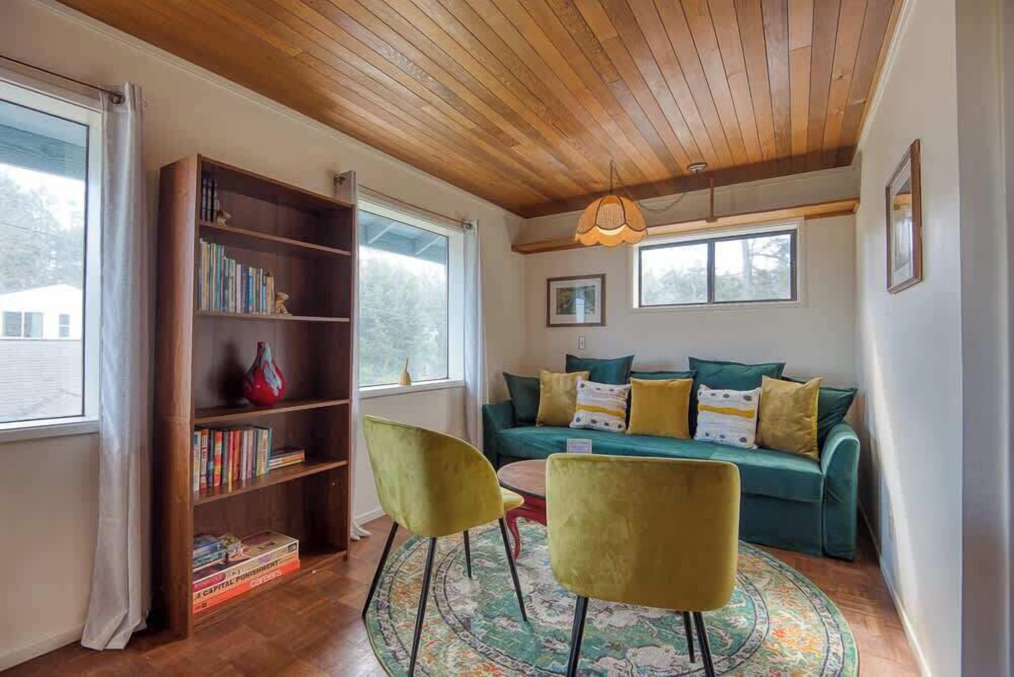 A cozy living room with a wooden ceiling, a green sofa with cushions, a bookshelf, two chairs, and a rug.