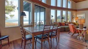 Een ruime woonkamer met een houten eettafel en stoelen, een bank, en grote ramen met uitzicht op het strand.