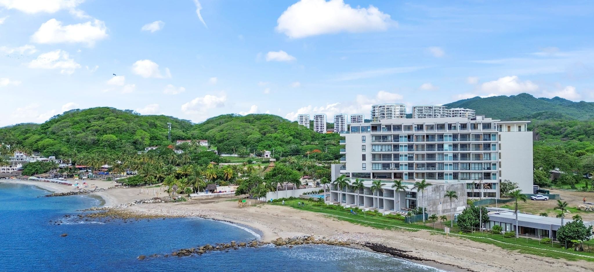 A beachfront hotel with multiple floors, a sandy beach, and lush greenery in the background.