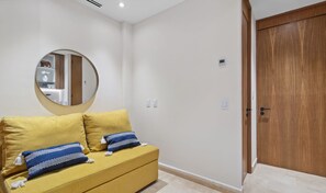 Quarto com sofá amarelo, espelho redondo, porta de madeira e parede branca.