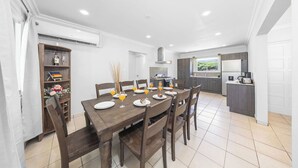 A dining area with a wooden table set for six, a kitchen in the background, and a view of greenery outside.