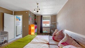 Un dormitorio con dos camas, una cuna, un radiador, una mesita y una ventana con cortinas.