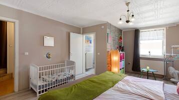A room with a crib, a bed, a radiator, a window with curtains, a wall-mounted shelf, and a ceiling fan.