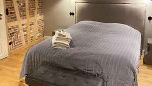 A bedroom with a large bed, a bench, a wall with exposed brick and wooden beams, and a nightstand with a lamp.