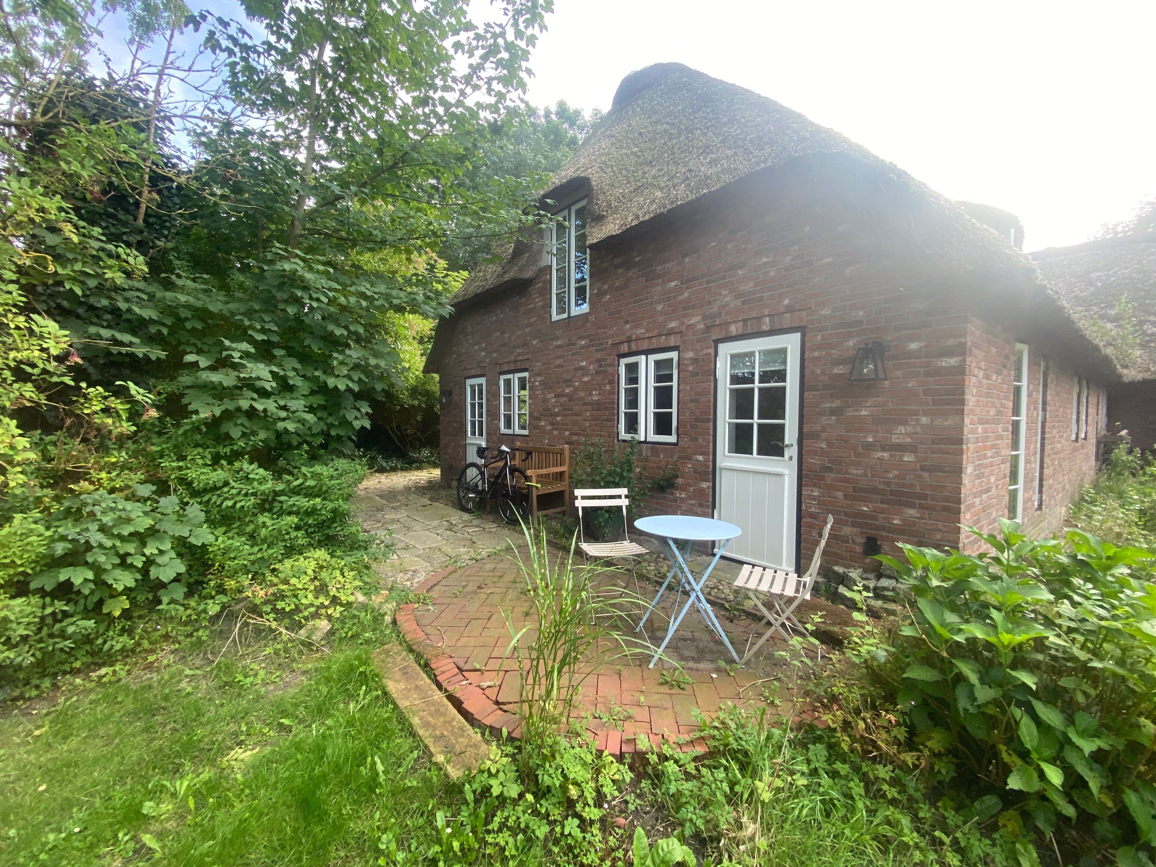 A brick house with a thatched roof, a garden with a paved path, and a table and chair set outside.