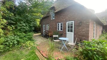 A brick house with a thatched roof, a garden with a paved path, and a table and chair set outside.