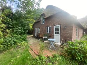 Una casa de ladrillo con techo de paja, un jardín con un sendero empedrado y un conjunto de mesa y sillas afuera.
