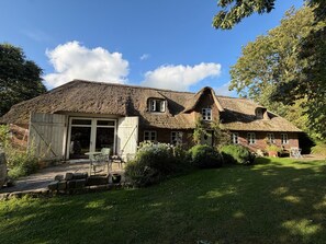 Une maison au toit de chaume, dotée d’un grand portail de garage, entourée d’une pelouse bien entretenue et d’arbres.