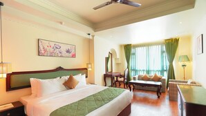 Une chambre d’hôtel avec un grand lit, un canapé, un bureau et une chaise. On y trouve un ventilateur de plafond, une fenêtre avec des rideaux et un tableau accroché au mur.