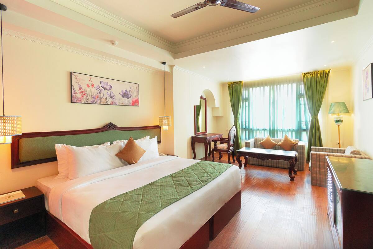A hotel room with a large bed, a sofa, a desk, and a chair. There is a ceiling fan, a window with curtains, and a painting on the wall.