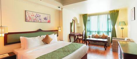 A hotel room with a large bed, a sofa, a desk, and a chair. There is a ceiling fan, a window with curtains, and a painting on the wall.