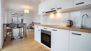 A modern kitchen with white cabinets, a wooden countertop, a built-in oven, and a stainless steel sink. There are two bar stools and a dining table in the adjacent dining area.