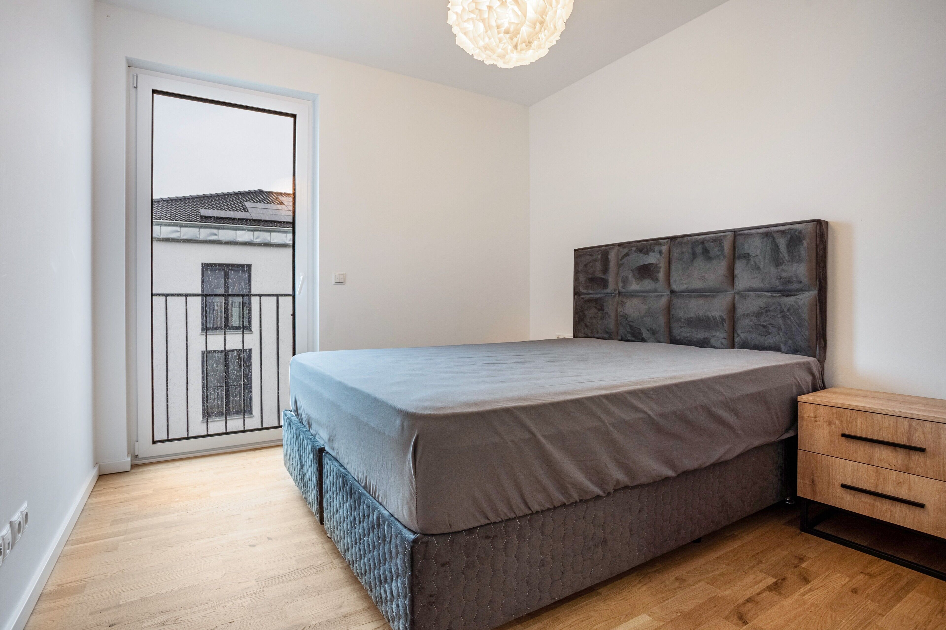 Une chambre à coucher comprenant un lit, une table de chevet en bois, une porte donnant sur un balcon et un luminaire au plafond.