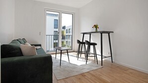 Een moderne woonkamer met een groene bank, een kleine tafel met stoelen en een uitzicht door een schuifdeur op een buitenbalkon.