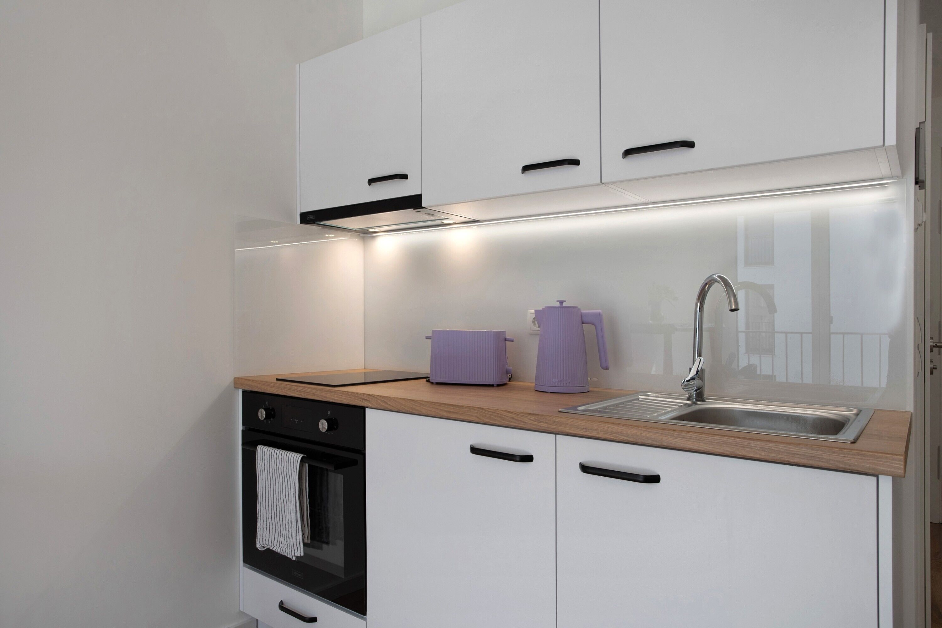Una cocina moderna con armarios blancos, encimera de madera, horno empotrado, fregadero y hervidor morado.