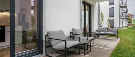 An outdoor patio area with a glass door leading to a kitchen, a seating area with two chairs and a table, and a small garden.