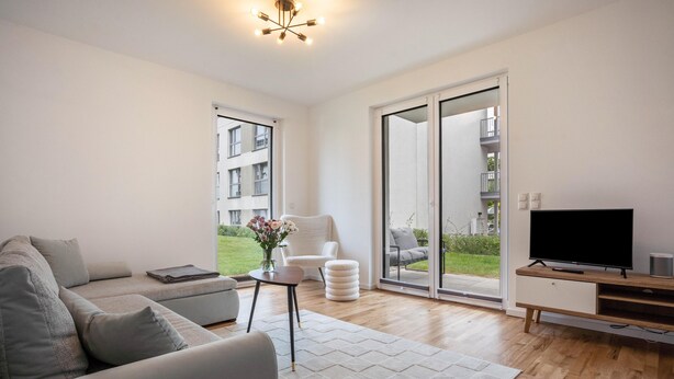 A modern living room with a sofa, a TV, a chair, a small table with a vase, and a sliding glass door leading to a balcony.